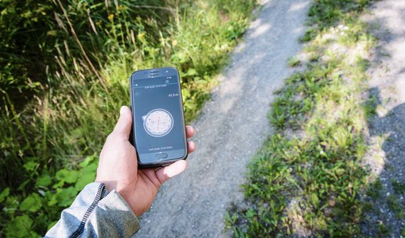 Bild: Eine Person, die auf ihrem Smartphone einen Kompass hat. Im Hintergrund ist ein Wanderpfad zu sehen.