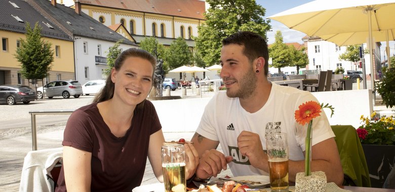 Bild: Ein Mann und eine Frau, die auf dem Marktplatz einen Brotzeitteller essen und Bier trinken. Im Hintergrund ist die Kirche zu sehen.