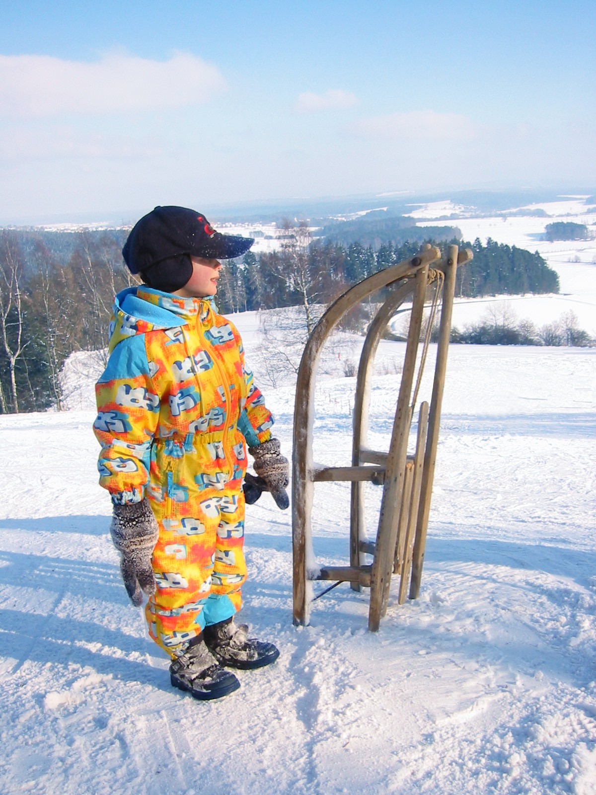 Bild: Ein kleiner Junge im gelben Schneeanzug, der vor seinem Schlitten im Winter steht.